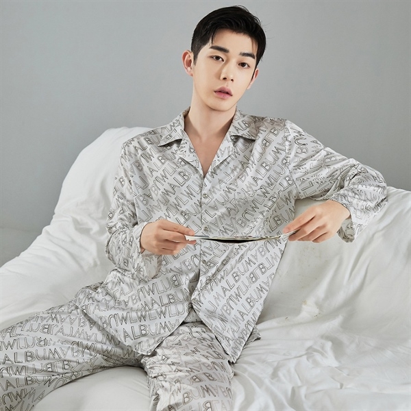 Bộ pijama lụa nam màu xám in chữ toàn thân, dáng dài tay quần dài, người mẫu ngồi trên giường trắng