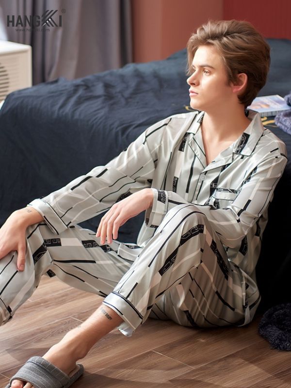 Nam giới mặc bộ pijama lụa kẻ sọc màu sáng ngồi trên sàn gỗ cạnh giường ngủ