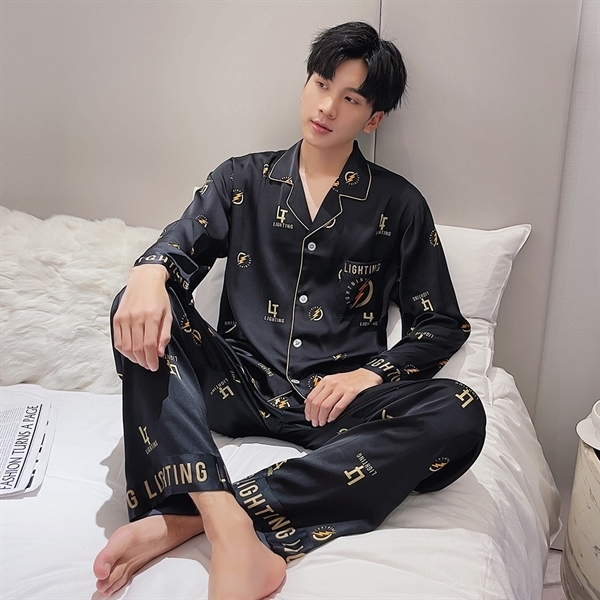 Nam giới mặc bộ pijama lụa đen tay dài họa tiết chữ vàng ngồi trên giường trắng